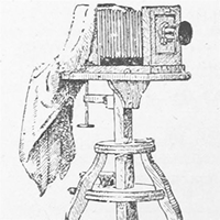 Illustration montrant un appareil photo posé sur un support en bois, couvert d'un tissu sombre.
